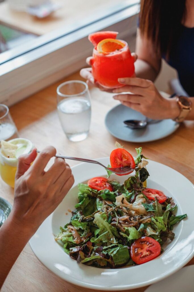 From above of unrecognizable person with fork eating delicious salad with tomatoes and lettuce while sitting in restaurant with anonymous friend